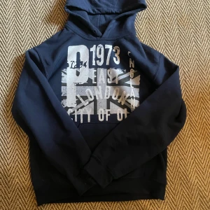 Mörkblå hoodie med tryck London - Snygg mörkblå hoodie med stort tryck på bröstet med texten '1973 East London City of Old' och motiv av Union Jack. Tröjan har huva, snörning och en klassisk . Perfekt för dig som gillar streetstyle och coola prints.