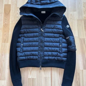 Svart pufferjacka från Moncler med huva - Svart pufferjacka från Moncler med stickade ärmar och ribbade muddar. Jackan har huva, dragkedja framtill och två fickor med knappstängning. Moncler-logga på ena ärmen och quiltad kropp i glansig polyester. Perfekt för kyliga dagar.