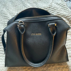 Svart handväska från Steve Madden - Snygg svart handväska från Steve Madden med stilren design och guldfärgad dragkedja. Väskan har två rejäla handtag, strukturerad yta och rymligt innerfack. Perfekt för dig som gillar klassisk look med en modern touch.