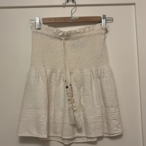 Vit kortkjol med snörning från Zara - Supersöt vit kortkjol från Zara med smockad midja, snörning med tofsar och dekorativa knappar framtill. Kjolen har volangkant och ett strukturerat tyg som ger en boho-vibe.