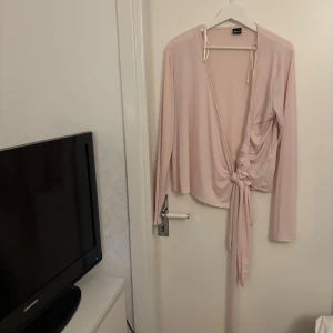 Ljusrosa omlottopp med knyt - Säljer en ljusrosa omlottopp från Gina tricot  med långa ärmar och djup v-ringning. Toppen har snygg knytning framtill och ett mjukt, tunt material som ger en skön känsla. Perfekt för dig som gillar trendiga och feminina plagg.