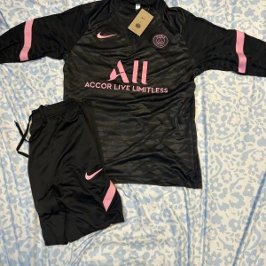 PSG svart långärmad fotbollströja Nike - Svart långärmad PSG fotbollströja från Nike med rosa detaljer och klubbmärke på bröstet. Tröjan har subtilt zebramönster i tyget och rosa logga samt text. Tillhörande svarta byxor med rosa detaljer och klubbmärke. Materialet är lätt och andas.