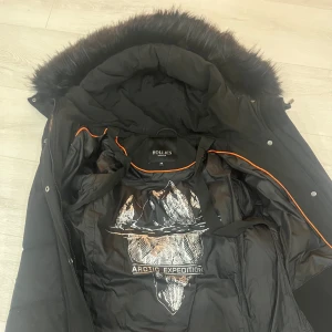 Svart lång pufferjacka från Hollies - Säljer en svart lång pufferjacka från Hollies med stor fluffig fuskpäls på huvan och snygga orange detaljer på insidan. Jackan har dragkedja, knappar i sidorna och är perfekt för kalla vinterdagar. Arctic Expedition-tryck på fodret ger extra cool känsla. Pris kan diskuteras 