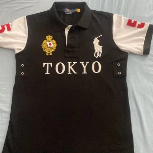 Polo Ralph Lauren Tokyo Chief Keef - Står storlek S men är stor i storlek. Svart och vit pikétröja från Polo Ralph Lauren med broderad Tokyo-text, guldigt emblem och stor vit polospelare på bröstet. Röda siffror på ärmar och rygg. Slim fit-modell med knappar framtill och korta ärmar. Perfekt för dig som gillar sportig och stilren look.