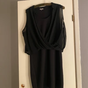 Svart omlottklänning från Truly Mine - Snygg svart kortklänning från Truly Mine med omlott-detalj framtill och ärmlös design. Klänningen har ett lager av tunn, transparent polyester över axlar och bröst, vilket ger en cool och modern look. Perfekt för dig som gillar stilrena och trendiga plagg.