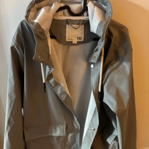 Grå regnjacka med huva från lager 157 - Snygg grå regnjacka från 157 med vit insida och huva med vita snören. Jackan har stora fickor framtill, tryckknappar och en stilren look. Perfekt för regniga dagar och enkel att matcha med allt. Materialet är syntet och jackan har lång ärm.