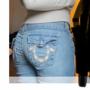 Ljusblå True Religion jeans bootcut - Säljer ett par ljusblå jeans från True Religion med klassisk orange brodyr på bakfickorna och snygga detaljer. Modellen har låg midja och bootcut-ben, tillverkade i mjukt jeanstyg. Perfekta för dig som gillar en avslappnad men trendig look. Långa på mig som är 165. Obs, Lite slitna längst ner