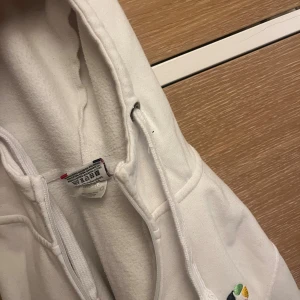 Vit hoodie med dragkedja Nordex - Vit hoodie från Nordex med broderad logga och text på bröstet. Tröjan har huva med snörning, dragkedja framtill och två fickor. Tillverkad i mjuk bomullsmix, perfekt för chill dagar eller när du vill vara bekväm.