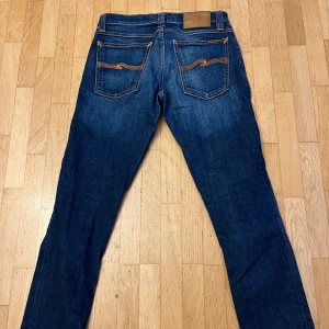 Mörkblå jeans från Nudie Jeans - Snygga mörkblå jeans från Nudie Jeans med klassiska orange sömmar och broderade bakfickor. Jeansen har en smal passform och är tillverkade i slitstarkt denim. Perfekta för dig som gillar en stilren och tidlös look.