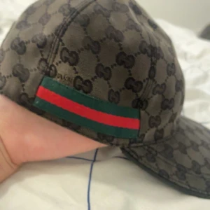 Gucci keps med GG-mönster och röd/grön detalj - Snygg keps från Gucci i mörkbrun färg med klassiskt GG-mönster över hela kepsen. På sidan finns en ikonisk röd och grön rand. Märket syns tydligt på insidan. Perfekt för dig som vill ha en lyxig och trendig accessoar.