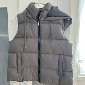 Grå dunväst med huva och dragkedja - Snygg grå dunväst med quiltad design och stor huva. Västen har dragkedja framtill, två fickor och justerbara snören vid huvan. Passformen är loose och materialet är syntet.