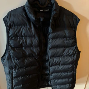 Svart dunväst från Zara - Snygg svart dunväst från Zara med quiltad design och hög krage. Västen har dragkedja framtill och två fickor på sidorna. Perfekt att slänga över en hoodie för en cool och avslappnad look.