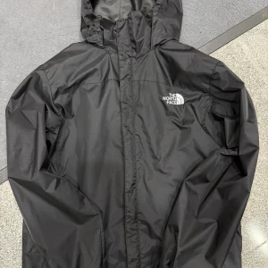 Svart vindjacka från The North Face - Svart vindjacka från The North Face med huva och logga på bröstet. Jackan har en stilren design med dragkedja framtill och är tillverkad i lätt polyester som står emot vind och regn. Perfekt för dig som gillar outdoor och streetstyle.