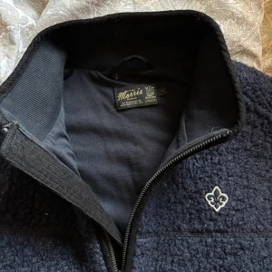 Morris mörkblå fleeceväst med dragkedja - Mysig mörkblå fleeceväst från Morris med broderad vit logga på bröstet. Västen har hög krage och hel dragkedja framtill. Perfekt att slänga på över en hoodie eller t-shirt för extra värme. Stilren och enkel design som funkar till många outfits. Pris diskuterbart vid snabb affär. Endast använd ett fåtal gånger.