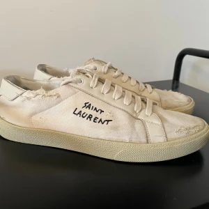 Saint Laurent vita sneakers - Säljer ett par vita Saint Laurent sneakers med handskriven logga på sidan. Skorna har råa, slitna kanter och detaljer i canvas och skinn. Klassisk rund tå, platt sula och snörning framtill. Perfekt för dig som gillar en edgy och avslappnad stil. Bra skick med mindre defekter som lite uppfläkt innuti, sulan har släppt pyttelite vid några få ställen och ett av slitdetaljerna har nått ner en bit extra men inget som direkt påverkar eller syns.🙌
