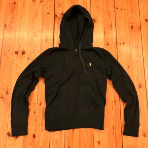 Svart hoodie från Polo Ralph Lauren - Svart hoodie från Polo Ralph Lauren med dragkedja, huva och snörning. Klassisk gul logga på bröstet och mjuk grå insida. Perfekt för chill dagar och enkel att matcha med jeans eller joggers.