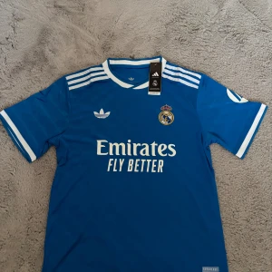 Blå Real Madrid x Adidas fotbollströja - Snygg blå Real Madrid fotbollströja från Adidas med vita detaljer och klassiska tre ränder på axlarna. Broderat klubbmärke på bröstet och Adidas Trefoil-logga. Tillverkad i lätt och ventilerande AEROREADY-material. Perfekt för dig som älskar fotboll och streetstyle.
