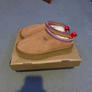 UGG slip-on mockasiner beige Tasman - UGG slip-on skor i beige mocka med mjukt foder och platt sula. Runt öppningen finns ett dekorativt band i rött och vitt samt en röd hällare bak. Perfekta för kalla dagar och riktigt sköna att glida runt i. Boxen förstörd pga den låg under sängen!