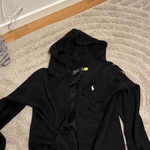 Svart hoodie från Polo Ralph Lauren - Svart hoodie från Polo Ralph Lauren med klassisk vit broderad logga på bröstet. Tröjan har huva med snörning och dragkedja framtill. Tillverkad i mjuk bomull, perfekt för en chill och stilren look. L passar som M