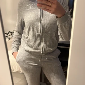 Mjaukis sett  - Grå hoodie från Juicy Couture med strassdetaljer på bröstet och ryggen. Tröjan har huva, långa ärmar, två fickor framtill och dragkedja. Mjuk och skön i bomullsmix, perfekt för chill dagar. Snygg och enkel att matcha med andra plagg. STORLEKEN ÄR 12/13 ÅR 