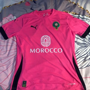 Marocko fotbollströja Puma rosa S - Rosa Marocko fotbollströja från Puma med svart logga och detaljer. Kortärmad modell i tekniskt material (RE:FIBRE), med Marockos landslagsemblem på bröstet och tryckt MOROCCO på framsidan. Perfekt för dig som vill sticka ut på planen eller läktaren.