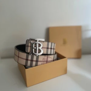 Burberry vändbart rutigt skärp - Snyggt vändbart bälte från Burberry med klassiskt rutigt mönster i beige, svart och brunt på ena sidan och svart rutigt mönster med präglad ryttare på andra sidan. Bältet har en silverfärgad metallspänne med Burberrys TB-logo. Tillverkat i skinn.