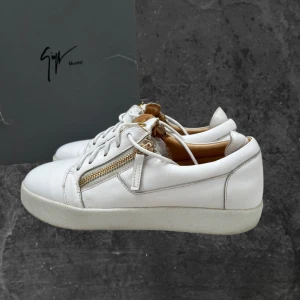 Vita sneakers från Giuseppe Zanotti - Snygga vita sneakers från Giuseppe Zanotti med guldfärgade dragkedjor och logga på plösen. Skorna är i skinn och har snörning samt platt sula. Perfekta för dig som gillar exklusiva detaljer och stilren design.