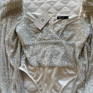 Glittrig body från Bella Barnett - Supersnygg body i silver från Bella Barnett med massor av paljetter som glittrar. Bodyn har långärmade puffärmar, v-ringning och figurnära passform. Perfekt för dig som vill sticka ut och glänsa på festen!