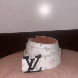 Vit bälte med monogram - Säljer ett snyggt vitt bälte med klassiskt monogrammönster och en svart spänne framtill. Bältet är gjort i skinn och har en stilren look som passar till många outfits.