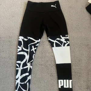 Svarta och vita träningsbyxor från Puma - Snygga leggings från Puma i svart med vita grafiska mönster och logga på benet. Hög midja och tight passform, perfekta för träning eller chill. Tillverkade i stretchigt syntetmaterial som sitter skönt och följer kroppen.