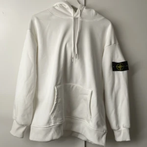 Vit hoodie från Stone Island - Säljer en clean vit hoodie från Stone Island med klassisk logga på ärmen. Tröjan har huva med snörning, stor magficka och ribbade muddar. Perfekt för dig som gillar streetwear och vill ha en stilren look.