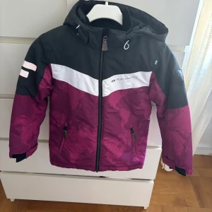 Mörkrosa vinterjacka från Lindberg - Snygg vinterjacka från Lindberg i mörkrosa och svart med vit detalj över bröstet. Jackan har kartmönster, huva, dragkedja och två fickor framtill. Perfekt för kalla dagar och har en sportig vibe. storlek 120