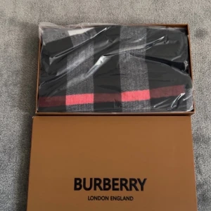 Burberry halsduk i kashmir - Exklusiv halsduk från Burberry i 100% kashmir med klassiskt rutigt mönster i svart, grått och rött. Broderad Burberry-logga och fransar i ändarna. Perfekt accessoar för att lyfta din outfit med en lyxig touch.