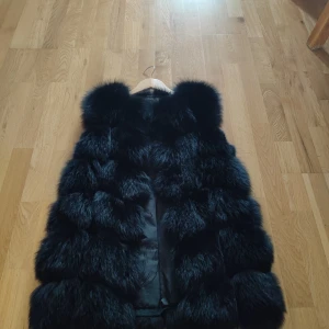 Svart fluffig pälsväst - Säljer en svart pälsväst med riktigt fluffig och voluminös look. Västen har breda, horisontella sektioner och är fodrad på insidan. Perfekt för dig som vill sticka ut och hålla värmen under kyliga dagar.