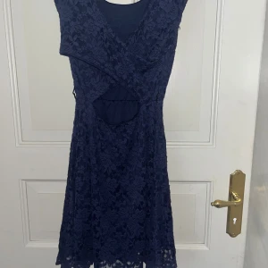 Mörkblå spetsklänning med cutout - Supersnygg mörkblå klänning i spets med v-ringning och cool cutout-detalj framtill. Klänningen är ärmlös och har en kort längd, perfekt för dig som gillar en feminin och trendig stil. Fodrad under spetsen för extra komfort.