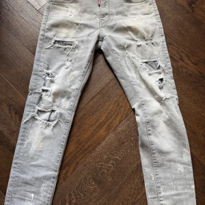 Grå ripped jeans från Dsquared2 - Snygga grå jeans från Dsquared2 med slitna och trasiga detaljer, färgstänk och coolt Dsquared2-märke bak. Modellen är slim och har klassisk femficksdesign. Skriv om du vill ha fler bilder och pris kan diskuteras. 