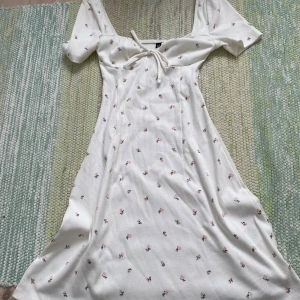 Vit blommig kortklänning från H&M Divided - Supersöt vit kortklänning från H&M Divided med små rosa blommor och ribbad struktur. Klänningen har korta ärmar, knytband vid bysten och en lätt utställd passform som ger en timglas ⌛️ figur . Perfekt för varma dagar och somriga looks!