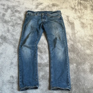 Levi's 511 blå jeans W32 L30 - Säljer ett par klassiska Levi's 511 jeans i blå tvätt med snyggt slitna detaljer. Modellen är slim fit med rak benform och normal midja. Jeansen har fem fickor, dragkedja och knappstängning. Perfekt för dig som gillar en tidlös och cool stil.