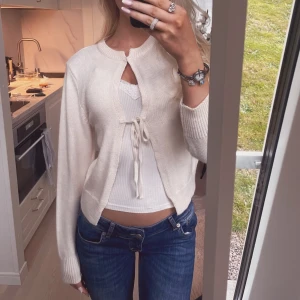 Beige stickad kofta med knyt detalj - Superfin beige stickad kofta med knyt framtill. Koftan har rund halsringning och långa ärmar, perfekt att slänga över ett linne eller topp. Enkel och stilren design som passar till jeans eller kjol.