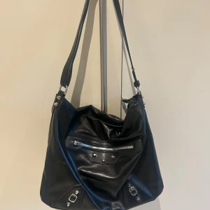 Svart axelväska i skinn med dragkedja - Snygg svart axelväska i mjukt skinn med justerbar rem och coola silverdetaljer. Väskan har en framficka med dragkedja och nitar, samt en rymlig insida. Perfekt för dig som vill ha en stilren och praktisk väska med lite edge.