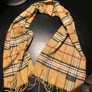 Rutig halsduk från Burberry i ull - HELT NY Klassisk rutig halsduk från Burberry i 100% lammull. Beige bas med svarta, vita och röda ränder, samt fransar i ändarna. Perfekt accessoar för att lyfta din outfit