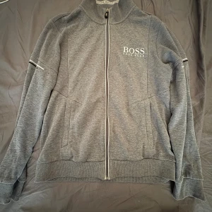 Grå ziptröja från Hugo Boss - Snygg grå ziptröja från Hugo Boss med vit logga på bröstet. Tröjan har hel dragkedja, ribbade muddar och detaljer på ärmarna. Perfekt för en clean och sportig look. Ståkrage och två fickor framtill.