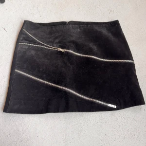 Svart mockakjol med dragkedjor från Att. - Cool svart kortkjol i mocka från Att. med två sneda silverfärgade dragkedjor framtill. Strl 34. Dragkedja bak. Midjemått ~78cm Längd ~33cm Bit av sömnen på sidan har gått upp men syns inte.