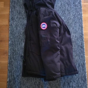 Svart dunjacka från Canada Goose - Säljer en svart dunjacka från Canada Goose med huva och klassisk logga på ärmen. Jackan är quiltad på insidan och har dragkedja samt knappar framtill. Perfekt för kalla vinterdagar och riktigt snygg streetstil.