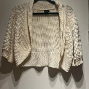 Bolero  - Säljer en krämvit stickad bolero från Lindex med trekvartsärmar och öppen front man kan även knäppa den. Koftan har grovstickad struktur och breda muddar, perfekt att slänga över en topp.