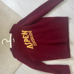 Vinröd Moncler stickad tröja Aspen - Vinröd stickad tröja från Moncler med gul text 'ASPEN COLORADO' på bröstet. Klassisk rund halsringning, ribbade muddar och lång ärm. Moncler-logga på ena ärmen. Perfekt för dig som gillar sportig och exklusiv stil.