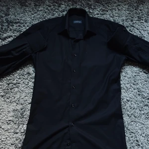 Svart skjorta från Tailor Store - Snygg svart skjorta från Tailor Store med klassisk krage och knäppning framtill. Skjortan är tillverkad i mjuk bomull med lite stretch för extra komfort. Perfekt för dig som gillar stilrena och enkla plagg.