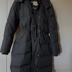 Svart lång dunjacka Boomerang S - Hej! Säljer min underbara jacka köpt ifrån Boomerang’s hemsida förra året. Varm och gosig med mycket fickor, dragsko i luvan och vid höfterna samt avtagbar luva.  Jag är 1,68 lång och den går ner en bit under knäna.   En pytteliten svag ”foundationfläck” finns på vänster sida utav jackan vid halspartiet. Se bild nummer 4. Ingenting som syns utåt alls, men påpekar ändå detta. Jackan är köpt för 7000:- nypris och jag säljer jackan för 4000:- med tanke på det fina skicket. Kom gärna m prisförslag! 