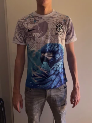 Nike Brasil Dri-Fit t-shirt med blå papegoja - Säljer en Nike Brasil Dri-Fit t-shirt i storlek L med ett sjukt snyggt tryck av en blå papegoja och blommor. T-shirten har korta ärmar, rund hals och är gjord i ett lätt och ventilerande polyestermaterial. Perfekt för dig som gillar unika och färgstarka plagg.