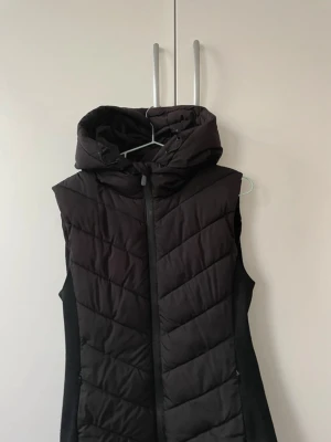 Svart quiltad dunväst med huva - Säljer en svart dunväst med stor, mysig huva och dragkedja framtill. Västen har ett quiltat mönster i diagonala ränder och är perfekt för lager-på-lager under kyliga dagar. Snygg och enkel att styla till olika outfits.